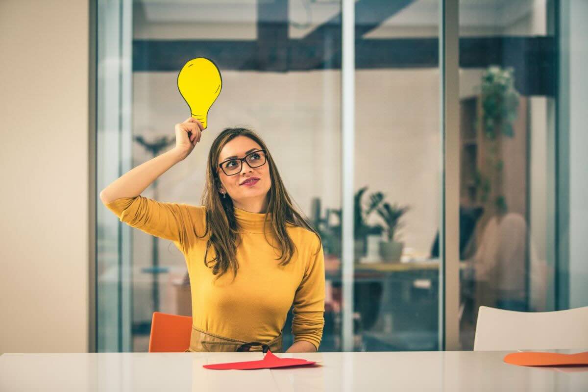 Ragazza vestita di giallo seduta al tavolo tiene in mano lampadina gialla in cartone